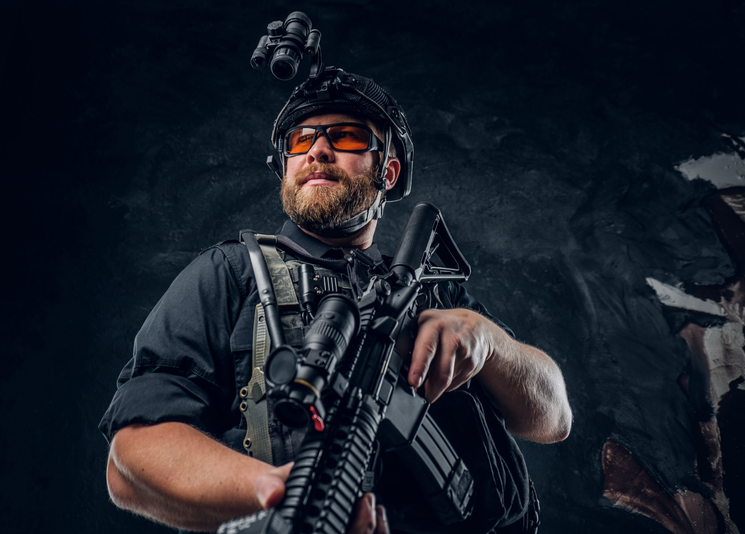 Bearded special forces soldier wearing body armor and helmet with night vision holding an assault rifle. Studio photo against a dark textured wall
